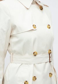 Krém színű ballonkabát gallérral, dupla soros kivitelben, nagy borostyánszínű gombokkal, övvel a derekán és oldalzsebekkel.