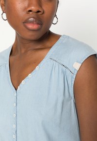 Lys blå denim topp med V-hals, dekorativ knappestolpe og kortermede med teksturdetaljer nær skulderen.