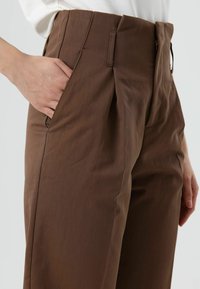 Hand gedeeltelijk in de zak van een high-waisted, bruine, getailleerde broek gedragen met een ingestopte witte blouse.