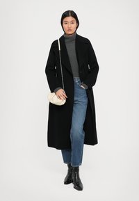 Un long manteau noir sur un col roulé gris, associé à un jean bleu et des bottines noires; tenant un sac couleur crème avec une bandoulière en chaîne.
