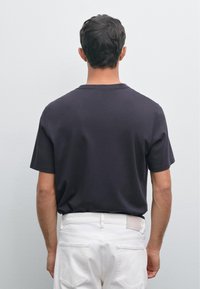 Uomo di fronte a un muro grigio chiaro, indossa una maglietta nera a maniche corte e pantaloni bianchi, in piedi con le braccia rilassate lungo i fianchi.