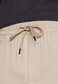 Beige joggerbyxor med dragsko i midjan med beige snören och svarta öglor, strukturerad tyg och sidofickor.