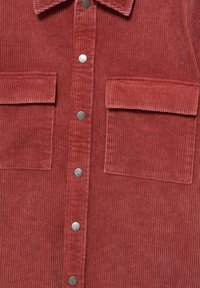 Chemise en velours côtelé rouge avec deux grandes poches à rabat sur le devant et des boutons-pression argentés au centre.