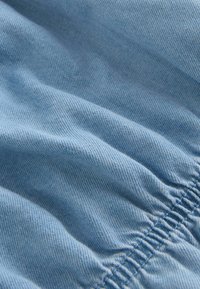 Tissu en denim bleu clair avec une texture lisse, présentant des fronces au bord inférieur et de subtiles lignes diagonales.