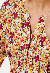 Bluse mit floralem Muster, das lebendige rote, gelbe und grüne Blumen auf weißem Hintergrund zeigt; Knopfdetails am Ausschnitt.