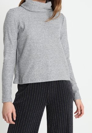 Frau trägt einen hellgrauen, gerippten Rollkragenpullover und dunkel gestreifte Hosen, steht vor einem schlichten weißen Hintergrund.