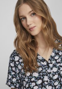 Blusa nera floreale con maniche corte, scollo a V, fiori bianchi e viola e bordi con bottoni, su uno sfondo a pattern discreto.