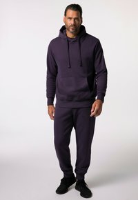 Fioletowa bluza z kapturem ze sznurkiem, kieszenią z przodu i pasującymi spodniami dresowymi. Tkanina wydaje się miękka i teksturowana, w zestawie z czarnymi butami treningowymi.
