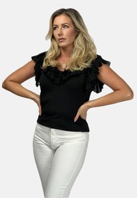 Czarny top z falbanami z dekoracyjnym dekoltem, w połączeniu z dopasowanymi białymi dżinsami. Prosty krój o gładkiej teksturze, podkreślający kontrast.
