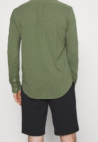 Mężczyzna w oliwkowej koszuli z długim rękawem i czarnych szortach, widziany z tyłu na jednolitym tle.