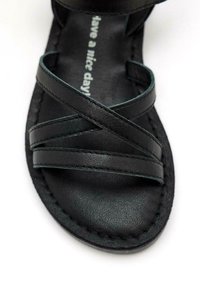 Sanda negra con múltiples tiras cruzadas de piel suave, con una plantilla de textura suave grabada con la frase "que tengas un buen día".