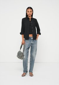 Camisa preta de botão sobre um top preto cropped, combinada com jeans claros desgastados e uma bolsa cinzenta texturada. Sandálias rasas pretas.