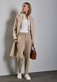 Beige wollen jas over een licht button-up shirt, witte tanktop en beige tapered broek. Witte sneakers en een bruine handtas maken de look compleet.