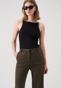 Black sleeveless top paired with olive green patterned trousers featuring white dots, belted waist, and pockets. Accessories include dark sunglasses.
