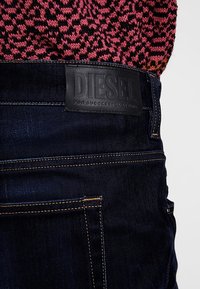 Tmavě modré džíny z denimu s kontrastním oranžovým prošíváním a texturovanou černou koženou nášivkou s vyraženým nápisem "DIESEL" na zadním pásku.