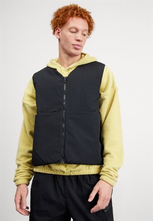 Gilet rembourré noir avec fermeture éclair à l'avant, poches surdimensionnées et finition texturée, porté par-dessus un sweat à capuche jaune clair.
