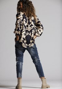 Bedruckte Bluse mit schwarz-beigem abstraktem Design, kombiniert mit zerrissenen Denim-Jeans und beigen Stiefeletten. Modell steht mit dem Rücken zur Kamera.