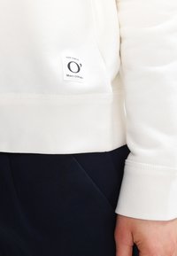 Witte sweatshirt met een gestikte zoom, geribde manchetten en een klein label met tekst. Gecombineerd met donkerblauwe stof. Soepele textuur.