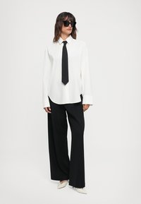 Camicia bianca a maniche lunghe con cravatta nera, abbinata a pantaloni neri larghi e scarpe bianche a punta, accessoriata con occhiali da sole neri.