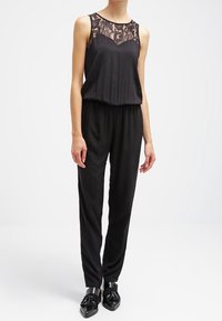Combinaison noire avec détail en dentelle au col, sans manches, taille élastique et pantalons fuselés, associée à des chaussures noires brillantes.