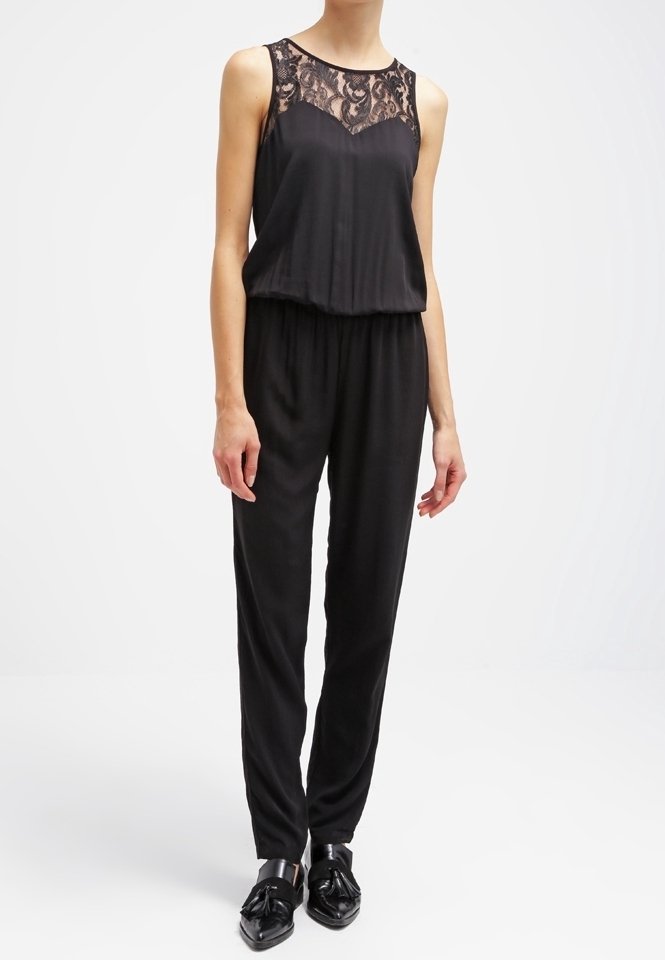 Combinaison noire avec détail en dentelle au col, sans manches, taille élastique et pantalons fuselés, associée à des chaussures noires brillantes.