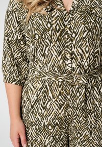 Chemise verte et crème avec un motif géométrique, dotée d'un col, de deux poches poitrine et d'une taille cintrée avec une boucle circulaire dorée.