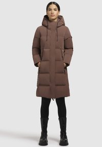 Bruine pufferjas met een capuchon, voorzien van een gestikt ontwerp, een ritssluiting aan de voorkant en zijzakken; gecombineerd met zwarte leggings en laarzen.