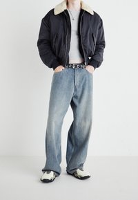 Chaqueta bomber negra con cuello de piel sintética beige, camisa ajustada gris, jeans sueltos de un azul claro y zapatillas negras y blancas.