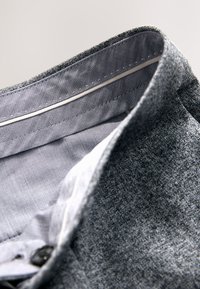 Gros plan du col d'une veste grise texturée montrant la couture de la doublure intérieure et le détail du bouton sur un fond clair.