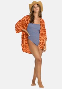 Kimono con estampado naranja sobre un traje de baño de una pieza a rayas azules y blancas. La modelo lleva un sombrero de paja y está descalza.