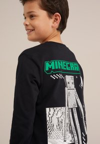 Fekete, hosszú ujjú póló zöld "MINECRAFT" logóval és a hátsó oldalon pixelált karaktergrafikával, puha pamut anyagból készült.