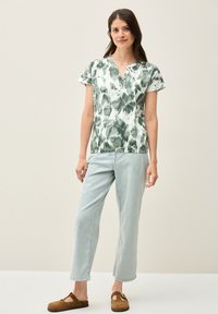 Top de tejido con estampado verde y blanco de mangas cortas, con escote en V y detalles de botones, combinado con pantalones vaqueros de mezclilla azul claro y zapatos marrones sin cordones.