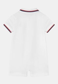 Bílý polo overal s krátkým rukávem, který má žebrovaný límec s červeným a námořnickým pruhováním a patentky na spodní části pro snadné oblékání.