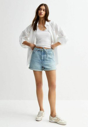 Shorts en denim bleu clair avec un ourlet retroussé, associés à un débardeur blanc et une chemise blanche surdimensionnée. Des baskets neutres complètent la tenue.