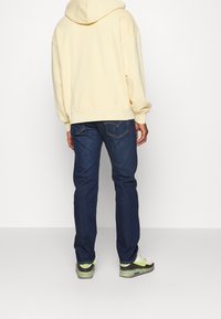 Sudadera con capucha de un amarillo claro y puños ajustados, combinada con jeans de un azul oscuro. Las zapatillas tienen una base blanca y detalles en verde neón.