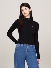 Donna con lunghi capelli scuri, che indossa un maglione nero a coste con zip e jeans blu a vita alta, in piedi con una mano sulla coscia.