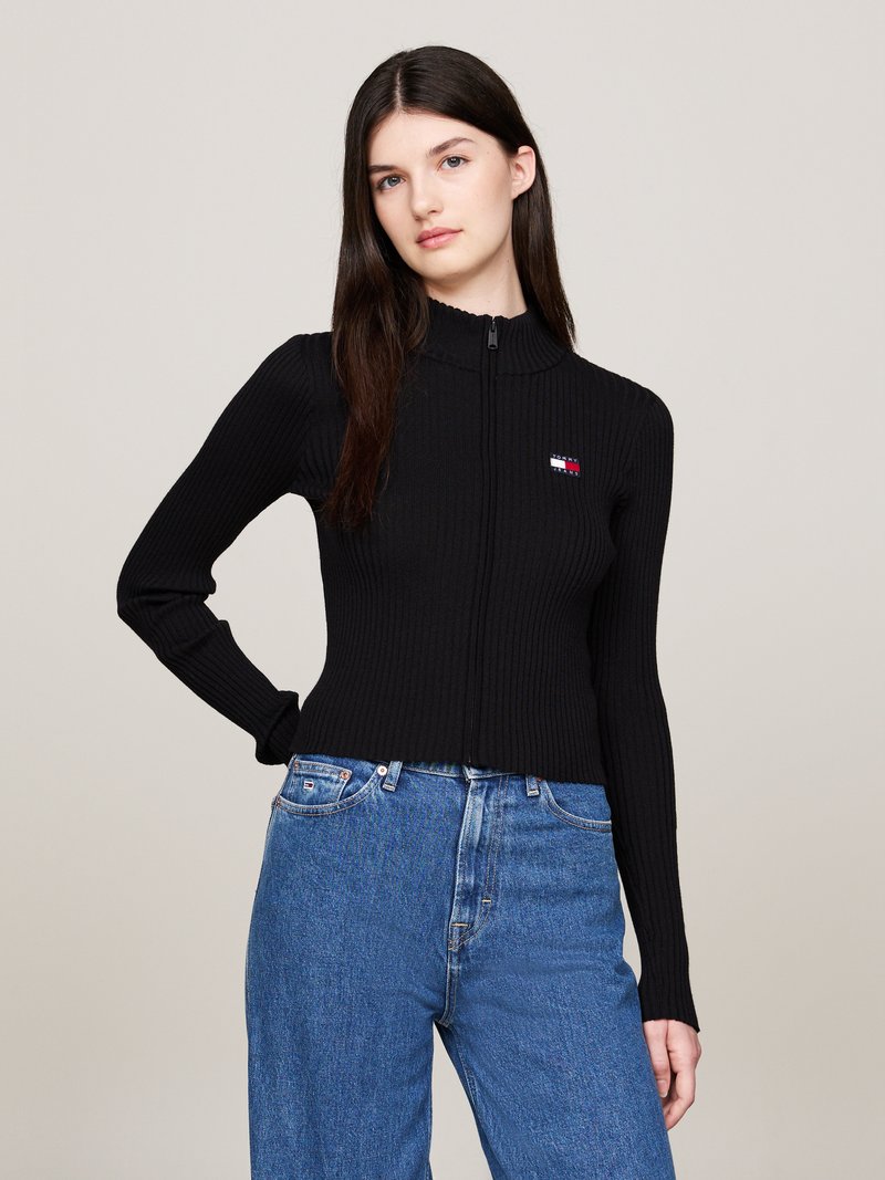 Donna con lunghi capelli scuri, che indossa un maglione nero a coste con zip e jeans blu a vita alta, in piedi con una mano sulla coscia.