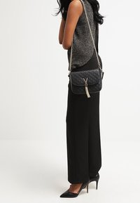 Kvinna i svarta vida byxor och ärmlös topp, bär svarta spetsiga klackskor och har en vadderad svart handväska med en guldfrans.