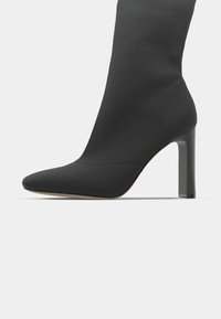 Stivaletto nero con tacco a stiletto, dal design elegante simile a una calza, punta affusolata, tessuto liscio e tacco alto a quadrato. Estetica minimalista.