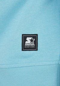 Ljusblått tyg med en strukturerad yta; har en svart fyrkantig patch märkt "STARTER BLACK LABEL" med en vit stjärnlogotyp.