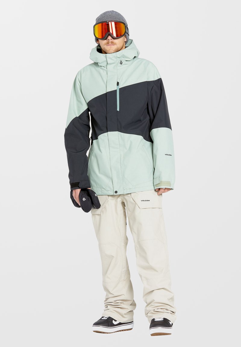 Chaqueta de snowboard en verde menta y negro, con cierre frontal, combinada con pantalones de snowboard beige y zapatillas negras con detalles blancos.