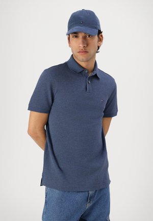 Tânăr purtând o șapcă albastră, tricou polo bleumarin și blugi albaștri, stând cu mâinile la spate pe un fundal simplu.