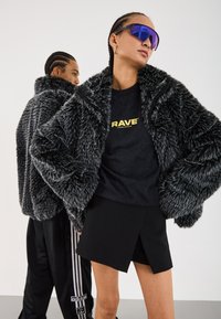 Zwei Personen tragen schwarze und graue texturierte Kunstpelzjacken; die Person vorne trägt lila Sonnenbrillen, ein schwarzes "RAVE"-Shirt und einen schwarzen Rock.