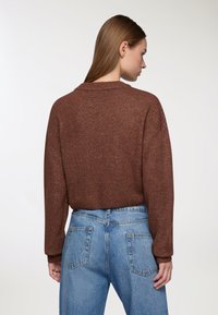 Donna con capelli castani lunghi e lisci, che indossa un maglione marrone e jeans blu a vita alta, in piedi di spalle contro uno sfondo neutro.