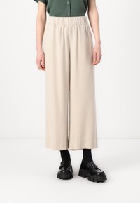 Beige vide benbukser med elastisk linning, laget av mykt stoff. Brukt med svarte kraftige loafers og mørke sokker.