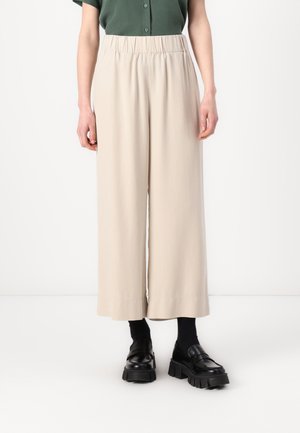 Pantaloni beige a gamba larga con vita elasticizzata, realizzati in un tessuto morbido. Indossati con mocassini neri chunky e calze scure.