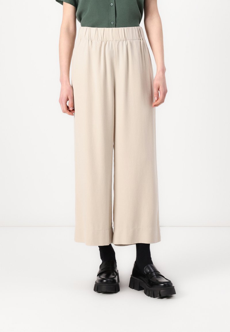 Beige vide benbukser med elastisk linning, laget av mykt stoff. Brukt med svarte kraftige loafers og mørke sokker.