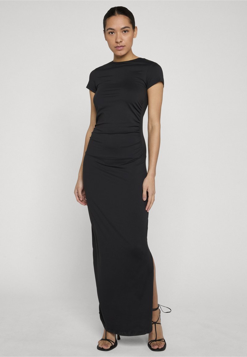 Femme aux cheveux foncés portant une robe maxi noire élégante à manches courtes et des talons noirs à lanières, debout devant un fond uni gris clair.