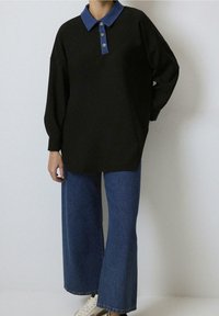 Pull noir oversize avec un col en denim bleu et une fermeture à trois boutons, associé à un jean bleu à jambes larges et à des baskets blanches.
