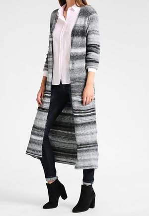 Kobieta ubrana w długi szary i czarny pasiasty kardigan na różowej koszuli, ciemne obcisłe jeansy i czarne botki, stojąca na białym tle.